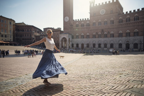 A woman in a sleeveless white top and a flowing blue skirt spins gracefully in the middle of Piazza del Campo in Siena, Italy, enjoying the lively atmosphere of the historic square. The brick-paved ground, arranged in a herringbone pattern, adds to the charm of the setting. In the background, the grand Palazzo Pubblico with its clock tower and arched windows stands tall, surrounded by groups of people walking and gathering.